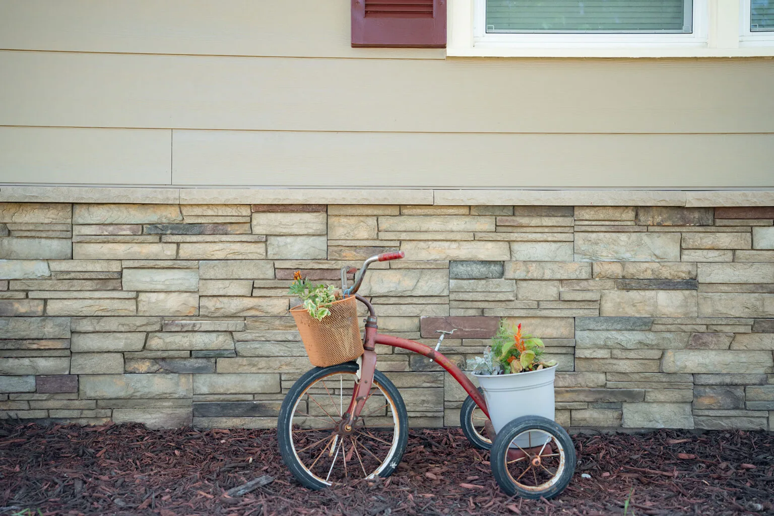 Close-up of beige fiber cement siding and stacked stone veneer installed by The Exterior Zone in Metamora, MI, with a vintage tricycle planter in the foreground.